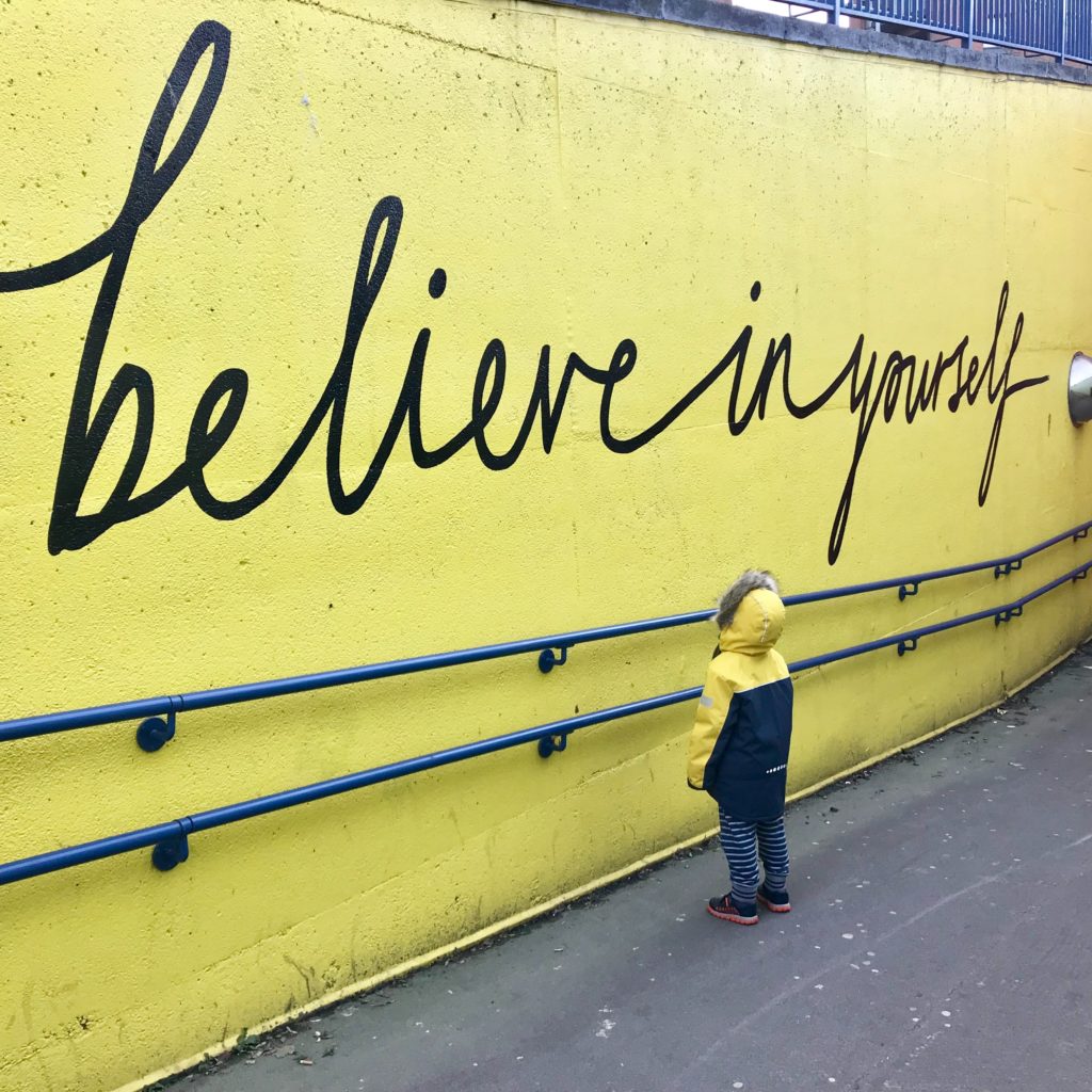 a child stands in front of a bright yellow wall that reads "believe in yourself"